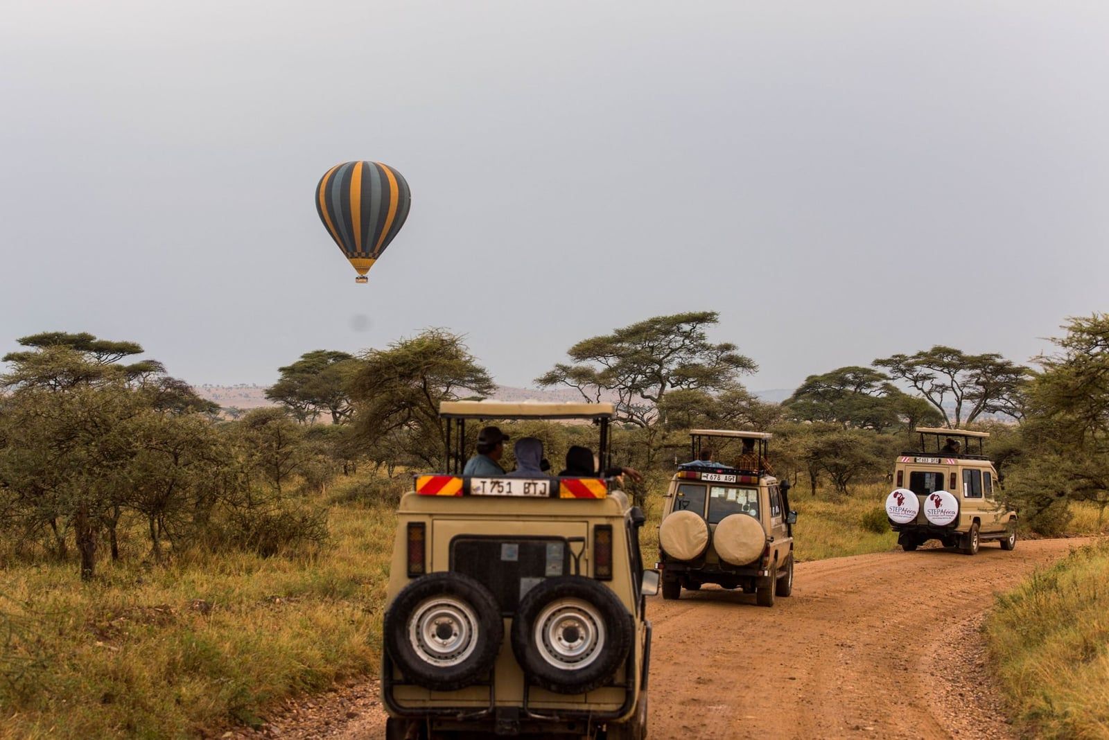 Veicoli da safari con tetti apribili percorrono una strada sterrata sotto una mongolfiera nel Parco Nazionale di Tarangire.